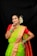 A woman dressed in a traditional green and orange saree adorned with gold jewelry, including necklaces, earrings, and a nose ring. She has her hair styled with white flowers and is wearing colorful bangles on her wrists. Her pose is confident and she is smiling.