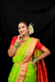 A woman dressed in a traditional green and orange saree adorned with gold jewelry, including necklaces, earrings, and a nose ring. She has her hair styled with white flowers and is wearing colorful bangles on her wrists. Her pose is confident and she is smiling.