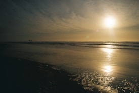 The image captures a serene beach scene during sunset. The sun is low on the horizon, casting a warm glow over the sea and reflecting on the wet sand. The sky is a mix of soft clouds and clear areas, enhancing the peaceful ambiance.