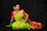 A woman in a traditional Indian saree poses gracefully against a dark background. She is adorned with elaborate jewelry, including necklaces, earrings, and bangles. Her saree is vibrant green with a rich red and gold border, complemented by a red blouse. Her hair is styled in an elegant updo, decorated with white flowers.