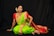 A woman in a traditional Indian saree poses gracefully against a dark background. She is adorned with elaborate jewelry, including necklaces, earrings, and bangles. Her saree is vibrant green with a rich red and gold border, complemented by a red blouse. Her hair is styled in an elegant updo, decorated with white flowers.