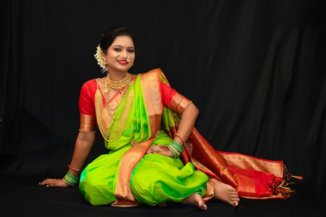 A woman in a traditional Indian saree poses gracefully against a dark background. She is adorned with elaborate jewelry, including necklaces, earrings, and bangles. Her saree is vibrant green with a rich red and gold border, complemented by a red blouse. Her hair is styled in an elegant updo, decorated with white flowers.