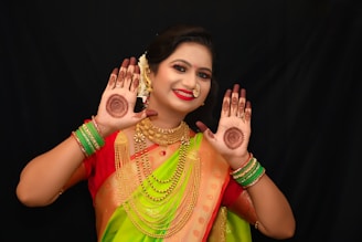 A woman is dressed in bright traditional attire with intricate jewelry. She has henna designs on her palms and is smiling, displaying the artwork. Her outfit includes a vibrant green and red sari, and she wears green bangles and gold-colored jewelry, including earrings and a necklace. Her hair is decorated with flowers.
