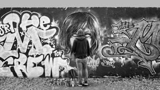 A person stands in front of a large graffiti-covered wall, holding a spray can. The graffiti includes large, bold text and a detailed illustration of a face with white eyes. There are several spray paint cans and a backpack on the ground near the person. The surroundings are littered with fallen leaves.