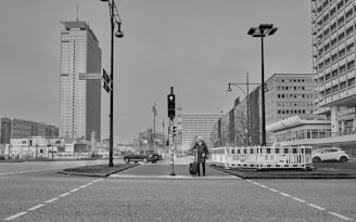 A black and white cityscape featuring a person with a suitcase at a crosswalk. Tall modern buildings line the street, with a prominent high-rise in the background. Streetlights and traffic signals are present, and a car is passing by.