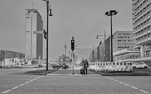 A black and white cityscape featuring a person with a suitcase at a crosswalk. Tall modern buildings line the street, with a prominent high-rise in the background. Streetlights and traffic signals are present, and a car is passing by.