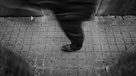 A person is shown walking with one leg in motion, creating a blurred effect. They are on a tiled ground with a pattern of square tiles. The scene is framed by concrete or stone walls on either side.