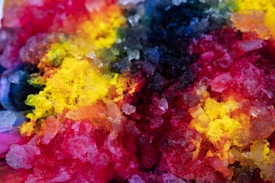 Crushed ice covered in vibrant and vividly colored syrups, creating a kaleidoscope of colors including red, yellow, blue, and black. The textures appear crystalline and translucent, with the colors blending and overlapping in some areas.