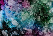 A close-up view of a colorful mixture of snow or crushed ice with hues of blue, green, purple, and pink. The texture of the ice appears jagged and crystalline, creating a vibrant and visually striking pattern. Underneath, hints of fabric with similar colors can be seen merging with the ice.