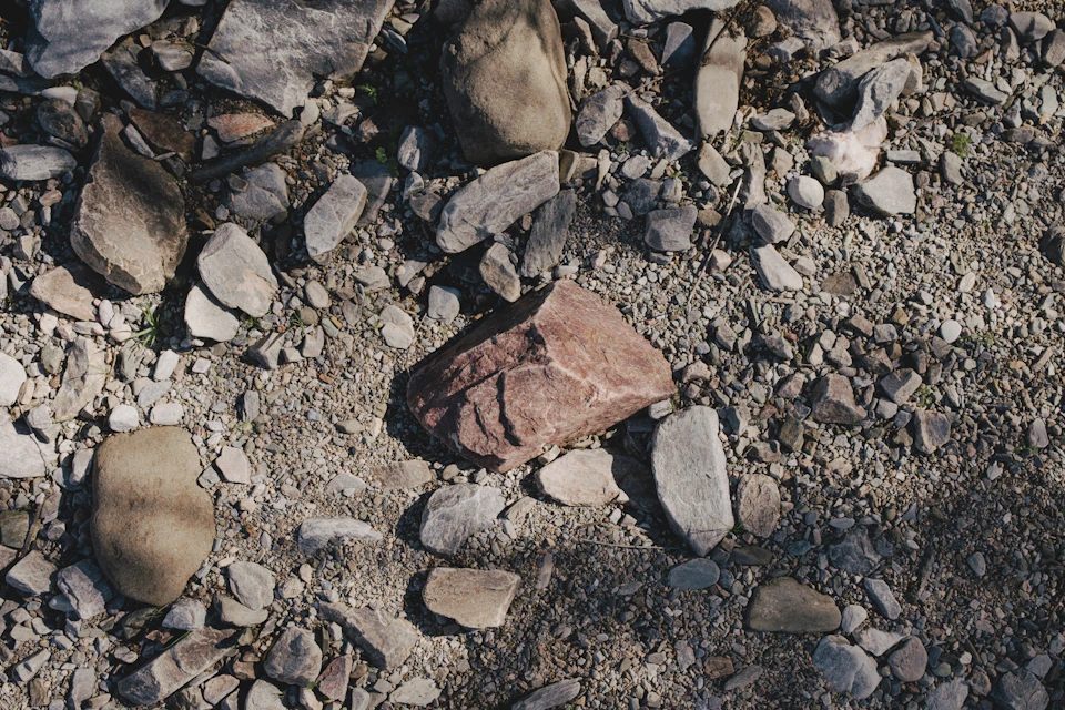 The image shows a rocky terrain with various types of rocks and boulders scattered across the ground, including large rocks, smaller stones, and larger boulders. The colors range from gray to brown, indicating different rock formations and textures. Some rocks are partially covered by dirt or other debris, adding depth to the scene. The perspective is from above, providing a bird's eye view of the landscape.