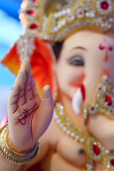 A close-up of a Ganesha idol, adorned with intricate jewelry, featuring detailed red and gold embellishments. The palm is raised in a blessing gesture with red markings and an artistic script. The facial features are gentle and benevolent, complemented by a golden crown.