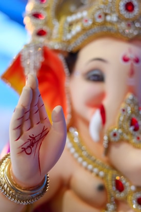 A close-up of a Ganesha idol, adorned with intricate jewelry, featuring detailed red and gold embellishments. The palm is raised in a blessing gesture with red markings and an artistic script. The facial features are gentle and benevolent, complemented by a golden crown.