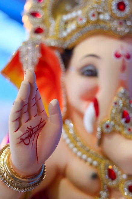 A close-up of a Ganesha idol, adorned with intricate jewelry, featuring detailed red and gold embellishments. The palm is raised in a blessing gesture with red markings and an artistic script. The facial features are gentle and benevolent, complemented by a golden crown.