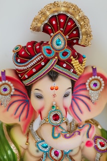 An intricately decorated statue of an elephant-headed figure adorned with vibrant jewels and ornate patterns. The headpiece is lavishly detailed with red fabric and gold embellishments, featuring bright blue and red gemstones. The figure has serene eyes, decorated ears, and an elaborate necklace, reflecting a rich cultural heritage.