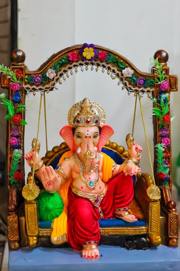 A colorful, intricately decorated statue of the Hindu deity Ganesha seated on an ornate swing adorned with flowers and jewels. The figure is dressed in bright red and gold attire, with multiple necklaces and a crown. The swing features detailed floral patterns and is embellished with vibrant colors.