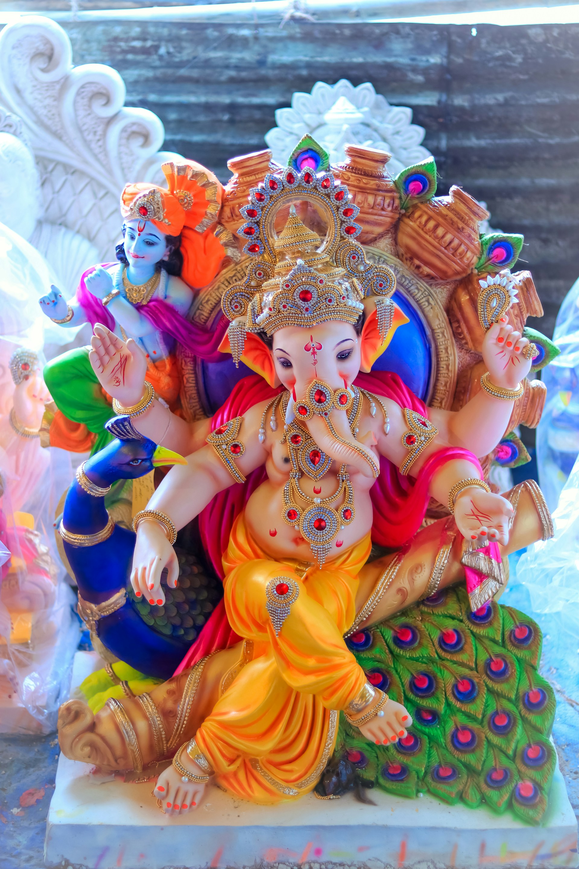 A colorful statue depicting deities, with ornate jewelry and vibrant clothing. The art piece has intricate details, including multiple arms, headdresses, and embellishments. A peacock sits at the base, adding to the richness of the design.