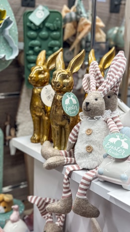 A display of Easter-themed decorations featuring plush bunnies with striped legs and gold figurine rabbits. The plush bunnies are dressed in whimsical outfits, sitting on a white shelf. They hold tags with 'Easter time' in pastel colors. The background includes additional decorative items like molds and soft textures.