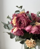A bouquet of dried flowers consisting of muted pink roses and beige carnations, accented with sage green leaves. The flowers have a vintage and rustic appearance, with petals that show subtle signs of withering.