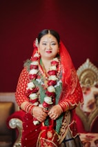 A woman is dressed in traditional attire with a red saree, adorned with intricate gold embroidery. She is wearing a garland of red and white roses around her neck and has henna on her hands. Her jewelry includes a headpiece and earrings, and her makeup highlights her features with vibrant red lipstick. The background features ornate furniture with a deep red and gold theme.