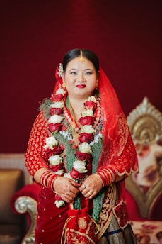 A woman is dressed in traditional attire with a red saree, adorned with intricate gold embroidery. She is wearing a garland of red and white roses around her neck and has henna on her hands. Her jewelry includes a headpiece and earrings, and her makeup highlights her features with vibrant red lipstick. The background features ornate furniture with a deep red and gold theme.