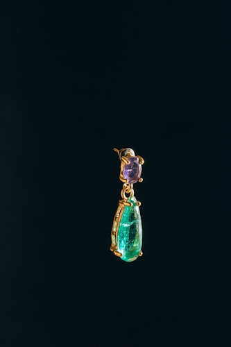 A single elegant earring with a purple gemstone at the top and a larger teardrop-shaped green emerald at the bottom. The gemstones are set in gold, and the earring is suspended against a dark, almost black background.