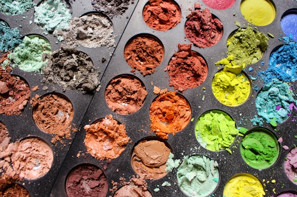 A close-up view of a palette filled with various colored powders. The powders are brightly colored, including shades of green, blue, brown, orange, red, and yellow. The texture of the powders is rough, some are spilled onto the palette, creating a vivid and chaotic pattern.