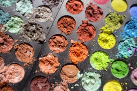 A close-up view of a palette filled with various colored powders. The powders are brightly colored, including shades of green, blue, brown, orange, red, and yellow. The texture of the powders is rough, some are spilled onto the palette, creating a vivid and chaotic pattern.