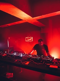 A DJ is performing in a dimly lit club or music venue, surrounded by DJ equipment including mixers and turntables. The room is bathed in an intense red light, creating a dramatic and vivid atmosphere. In the background, a neon sign adds to the ambiance.