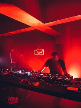 A DJ is performing in a dimly lit club or music venue, surrounded by DJ equipment including mixers and turntables. The room is bathed in an intense red light, creating a dramatic and vivid atmosphere. In the background, a neon sign adds to the ambiance.