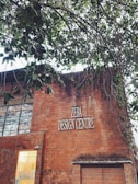 A brick building with a sign reading 'ZEBA DESIGN CENTRE'. Part of the building’s facade includes large windows and a smaller section with a lit interior visible through a glass panel. Above the building, tree branches with green leaves extend across the upper part of the image.