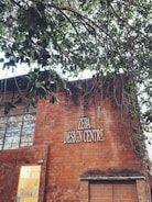 A brick building with a sign reading 'ZEBA DESIGN CENTRE'. Part of the building’s facade includes large windows and a smaller section with a lit interior visible through a glass panel. Above the building, tree branches with green leaves extend across the upper part of the image.