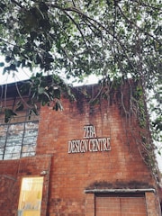 A brick building with a sign reading 'ZEBA DESIGN CENTRE'. Part of the building’s facade includes large windows and a smaller section with a lit interior visible through a glass panel. Above the building, tree branches with green leaves extend across the upper part of the image.
