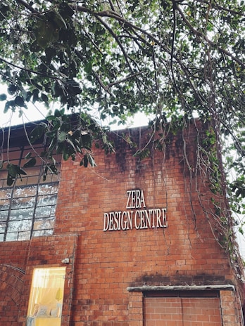 A brick building with a sign reading 'ZEBA DESIGN CENTRE'. Part of the building’s facade includes large windows and a smaller section with a lit interior visible through a glass panel. Above the building, tree branches with green leaves extend across the upper part of the image.