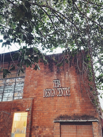 A brick building with a sign reading 'ZEBA DESIGN CENTRE'. Part of the building&rsquo;s facade includes large windows and a smaller section with a lit interior visible through a glass panel. Above the building, tree branches with green leaves extend across the upper part of the image.