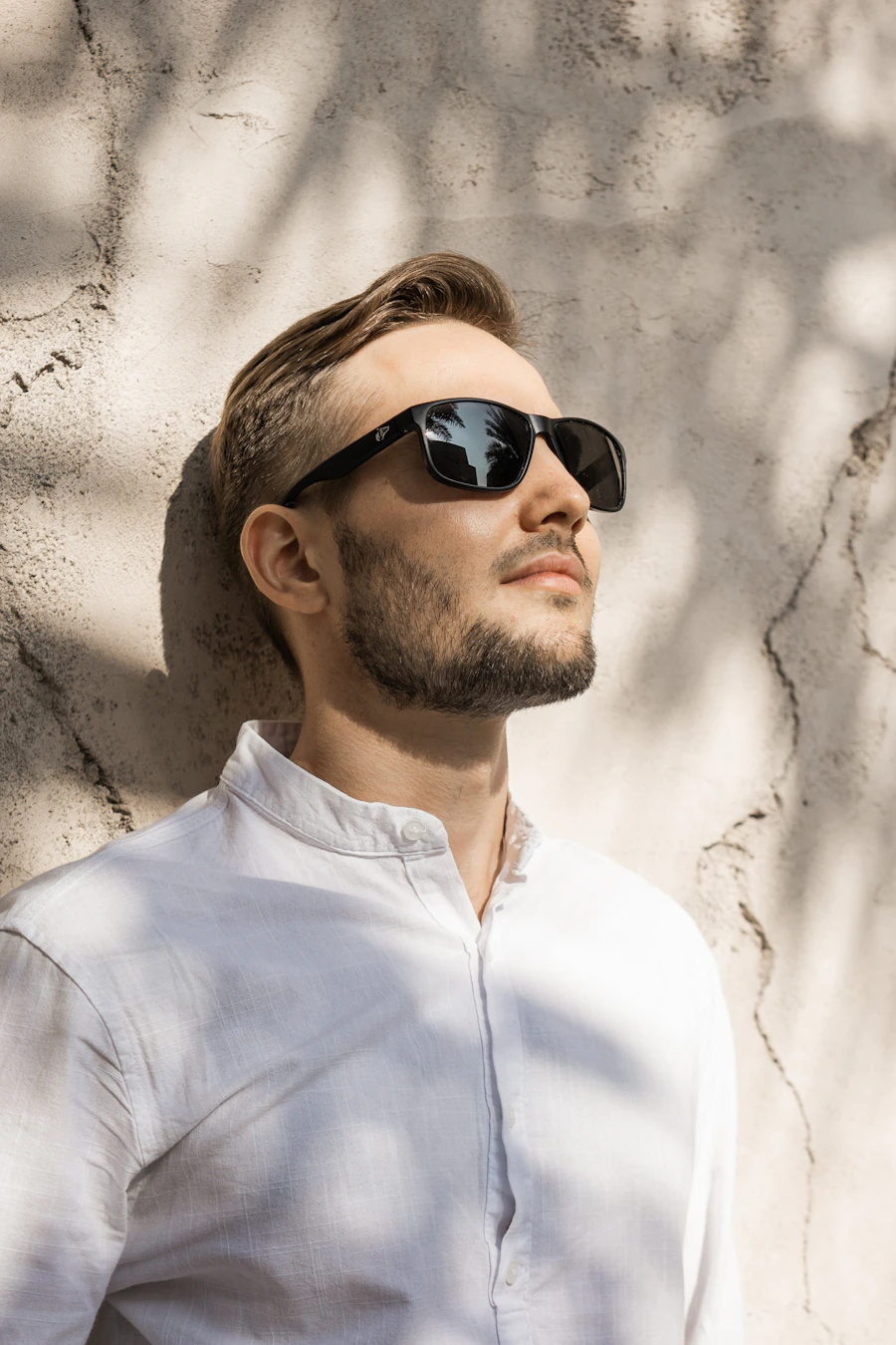 Man in sunglasses and a relaxed shirt leaning on a pale wall