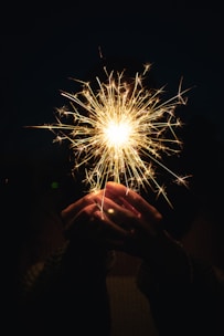 Hands holding a lit sparkler with glowing sparks around.