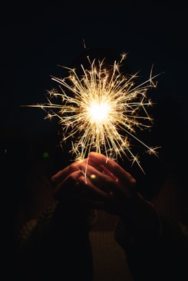 Hands holding a lit sparkler with glowing sparks around.