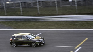 A black compact car is driving on a race track against a blurred green and metal railing background, indicating motion. The car is sleek with reflective surfaces, and the track is marked with white and yellow lines.