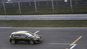 A black compact car is driving on a race track against a blurred green and metal railing background, indicating motion. The car is sleek with reflective surfaces, and the track is marked with white and yellow lines.