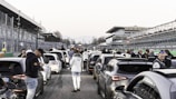 A crowded race track with several high-performance cars lined up. People are gathered around, some engaging in conversations or checking their phones. The atmosphere is bustling, with a spectator stand on one side and several visible structures in the background.