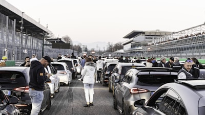 A crowded race track with several high-performance cars lined up. People are gathered around, some engaging in conversations or checking their phones. The atmosphere is bustling, with a spectator stand on one side and several visible structures in the background.