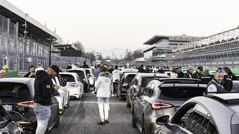 A crowded race track with several high-performance cars lined up. People are gathered around, some engaging in conversations or checking their phones. The atmosphere is bustling, with a spectator stand on one side and several visible structures in the background.