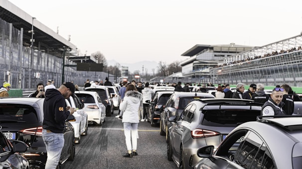 A group of legends cars lined up on a racetrack with enthusiastic drivers and fans in the background.