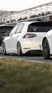 A white car with sporty modifications is prominently displayed, featuring sleek lines and large alloy wheels. It is parked on an asphalt surface, with other vehicles lined up nearby and blurred figures in the background, suggesting a car meet or event. The car's distinctive rear lights and exhaust pipes add to its dynamic appearance.
