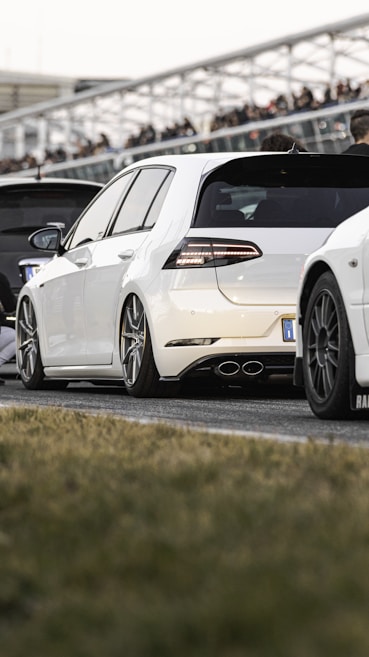 A white car with sporty modifications is prominently displayed, featuring sleek lines and large alloy wheels. It is parked on an asphalt surface, with other vehicles lined up nearby and blurred figures in the background, suggesting a car meet or event. The car's distinctive rear lights and exhaust pipes add to its dynamic appearance.