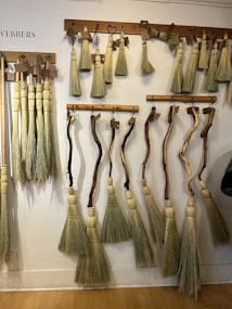 Several handmade brooms with wooden handles and straw bristles are hung on display racks against a white wall. The brooms vary in shape, featuring both straight and curved handles. Each broom is crafted with attention to detail, and some have decorative elements. Labels are attached to many of the brooms.