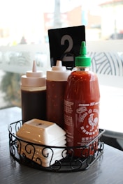 A wire basket on a table contains three bottles of condiments, including a prominent bottle of Sriracha hot sauce with a green cap. There is a black table number card displaying the number 2. The setting appears to be a casual dining restaurant with frosted glass windows in the background.