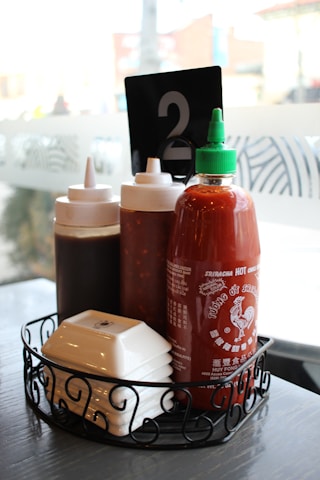 A wire basket on a table contains three bottles of condiments, including a prominent bottle of Sriracha hot sauce with a green cap. There is a black table number card displaying the number 2. The setting appears to be a casual dining restaurant with frosted glass windows in the background.