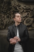 A person wearing a black leather jacket and a white shirt stands against a detailed, ornate backdrop featuring intricate leaf and flower carvings. The individual appears to be adjusting their sleeve while looking into the distance.
