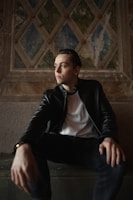 A person is seated in a contemplative pose against a decorative, textured wall featuring a geometric pattern with muted colors. The individual wears a black leather jacket over a white shirt and dark pants, with their gaze directed slightly to the side, creating a thoughtful expression.
